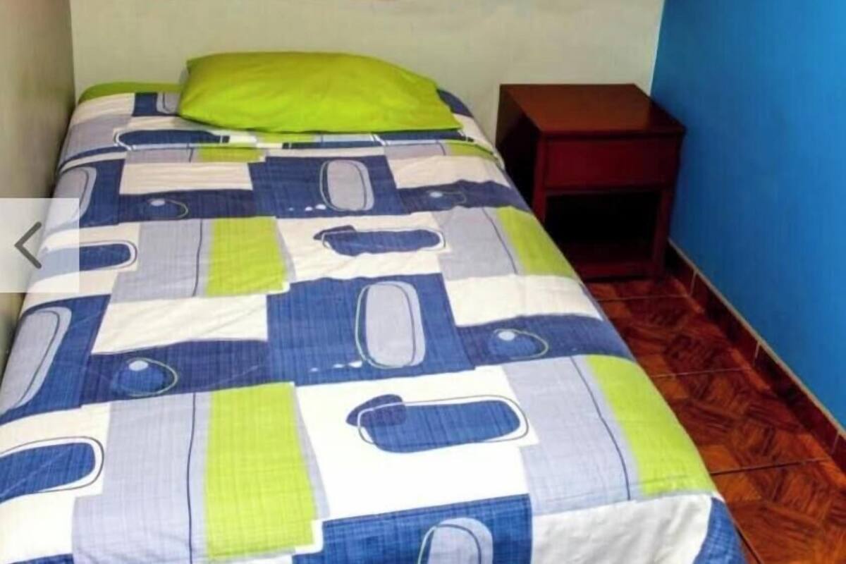 A single bed with a patterned bedspread, a red curtain, and a small wooden nightstand.