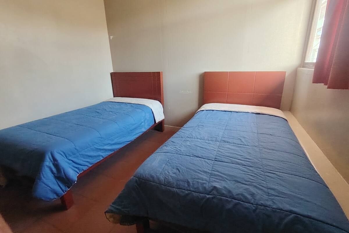 Two single beds with wooden headboards and blue bedding in a room with plain walls and a window with curtains.