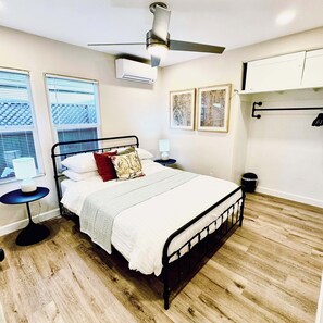 Een slaapkamer met een bed, een plafondventilator, een airconditioner, een nachtkastje met een lamp, een raam met jaloezieën en een wandplank.