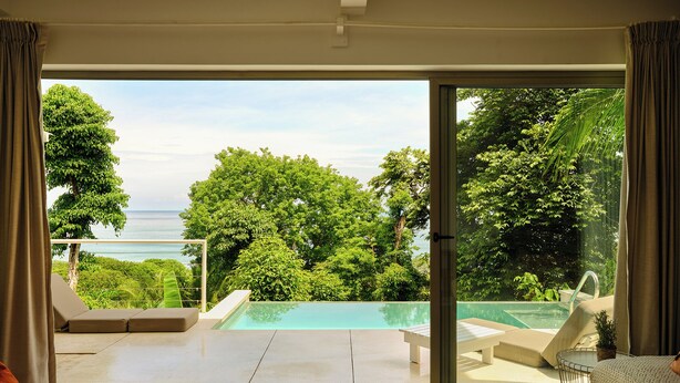 Uma sala de estar moderna com uma ampla porta de vidro deslizante que dá para uma área de piscina privativa, com vista para o oceano e vegetação exuberante.