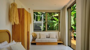Quarto com uma cama grande, guarda-roupa de madeira, um banco com almofadas e vista para um verde exuberante pela janela.