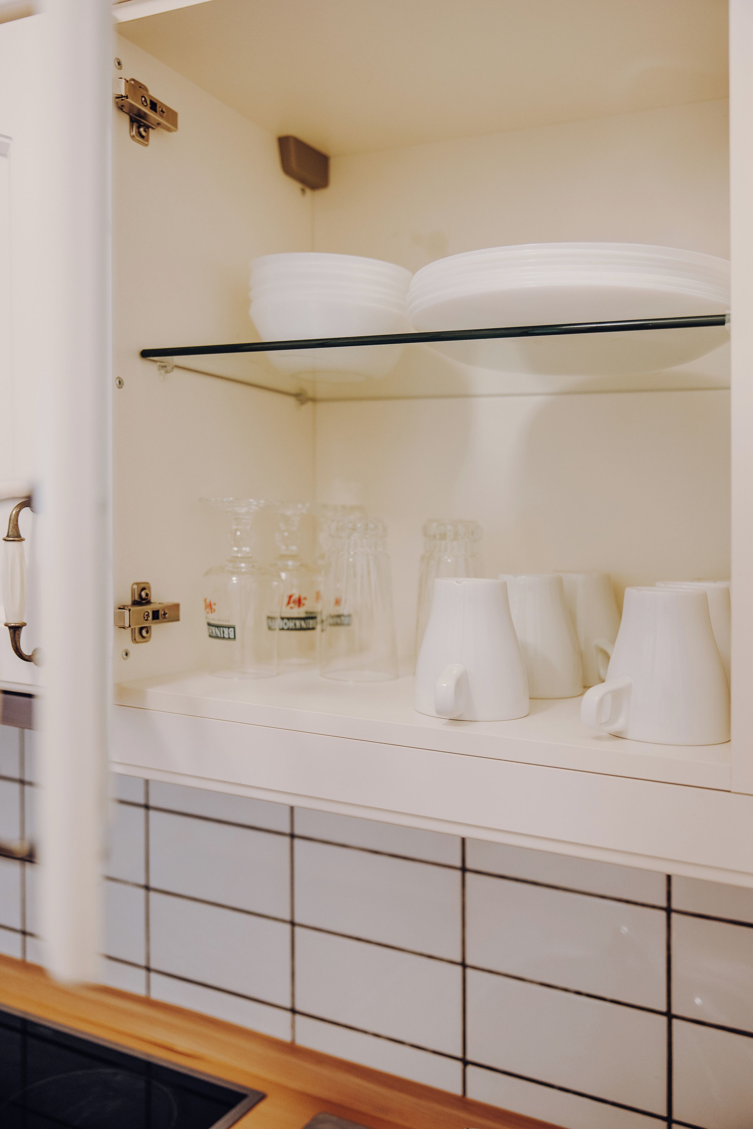 Una cocina con armarios blancos, salpicadero de azulejos y una repisa de vidrio con vajilla y cristalería blancos.