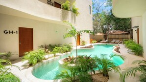 Zona de piscina con diseño curvo, rodeada de abundante vegetación y un edificio con balcones.