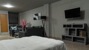 Ein Hotelzimmer mit einem großen Bett, einem an der Wand montierten Fernseher, einer kleinen Sitzecke mit Sofa und Sesseln sowie einer Küchenzeile im Hintergrund.