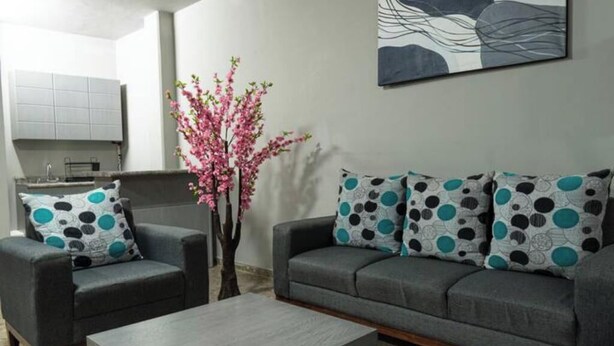 Ein Wohnzimmer mit grauem Sofa, zwei Sesseln und einem Couchtisch. Ein Blumenvasen mit rosa Blumen und ein abstraktes Kunstwerk an der Wand sind ebenfalls zu sehen.