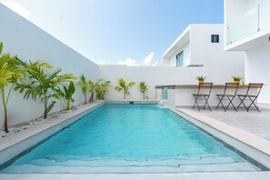 Une piscine extérieure moderne, avec un mur blanc et des plantes en pot, entourée d’un bâtiment blanc.