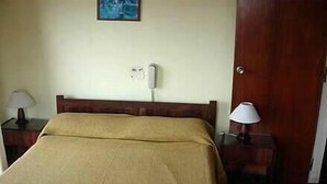 Uma cama de solteiro com cabeceira de madeira, mesa de cabeceira com abajur e telefone fixado na parede.