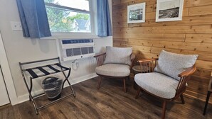 Une pièce aux murs en bois, avec deux fauteuils rembourrés, une fenêtre avec des rideaux, un radiateur et une petite étagère sur laquelle se trouve une boîte.