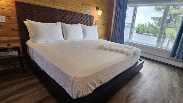 Un lit avec du linge de lit blanc, une tête de lit en bois et une vue sur une étendue d’eau par la fenêtre.