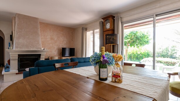 A living room with a wooden table, a sofa, a fireplace, and a large window with a view of greenery.