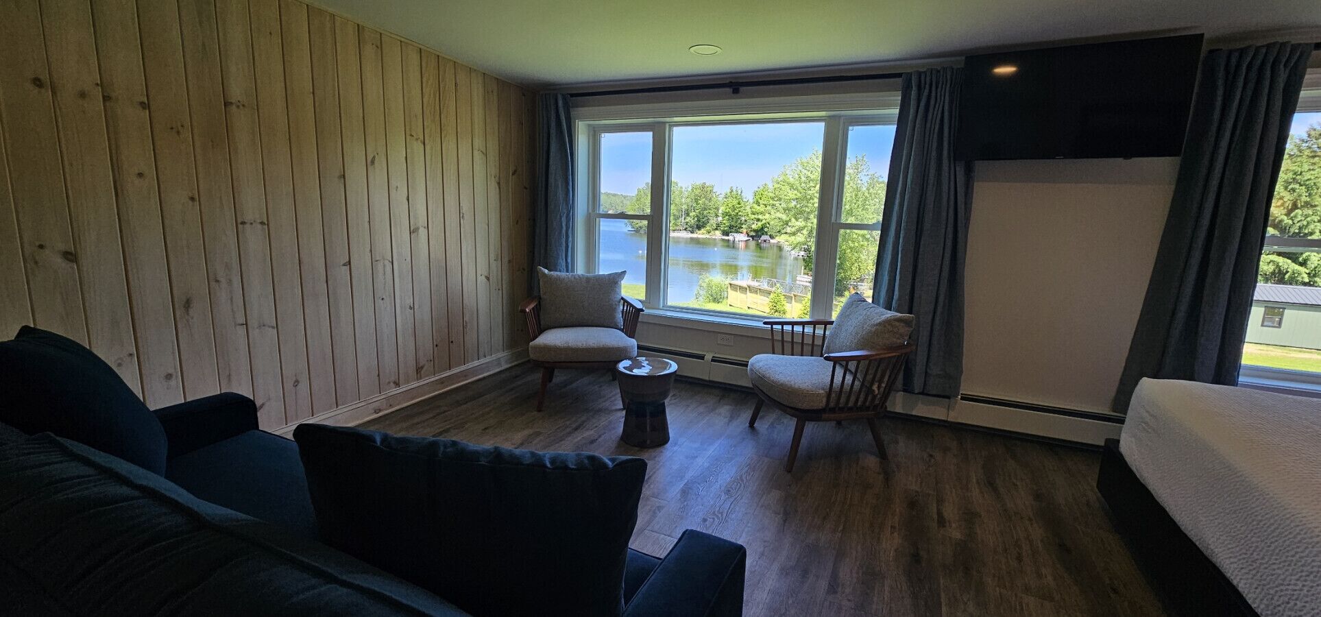 Een kamer met houten vloer, een groot raam met uitzicht op een meer, twee fauteuils, een kleine ronde tafel en een bed.