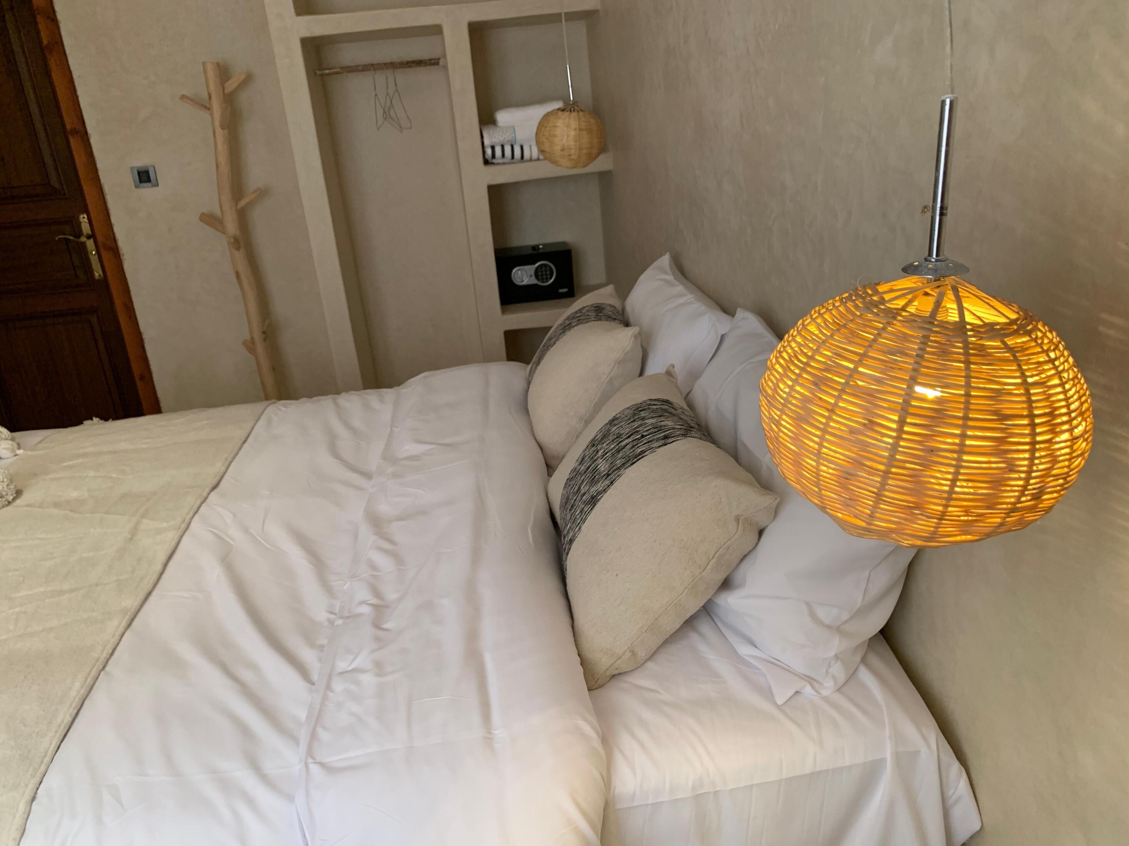 Een opgemaakt bed met wit beddengoed en meerdere kussens, een bedlampje, en een plank met een mand en een camera.