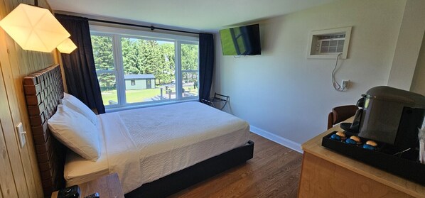 A hotel room with a bed, a TV mounted on the wall, a coffee maker, and a window with a view of the outdoors.