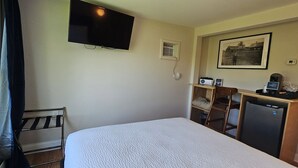 A hotel room with a bed, a TV mounted on the wall, a small desk with a coffee maker, a chair, and a picture frame on the wall.
