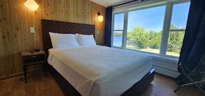 Ein Schlafzimmer mit einem großen Bett, Holzwänden, einem Nachttisch, einer Lampe und einem Fenster mit Blick auf Bäume und einen See.