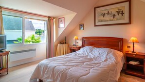A bedroom with a bed, a TV, a window with curtains, a painting, and a lamp.