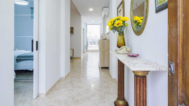 Un pasillo con suelo de mármol, una puerta de madera, una consola de mármol con un jarrón de girasoles y un espejo con marco floral.