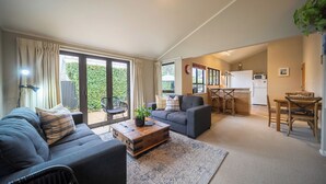 A living room with a sofa, armchair, and a coffee table. There is a sliding glass door leading to a balcony with a view of greenery. A kitchen area is visible in the background. - Townhouse Retreat - Te Anau Getaway (Te Anau)