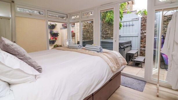 A bedroom with a bed, a small table, and a chair. There is a window and a door leading to an outdoor area with a hot tub. - Sea Sanctuary (Bideford)