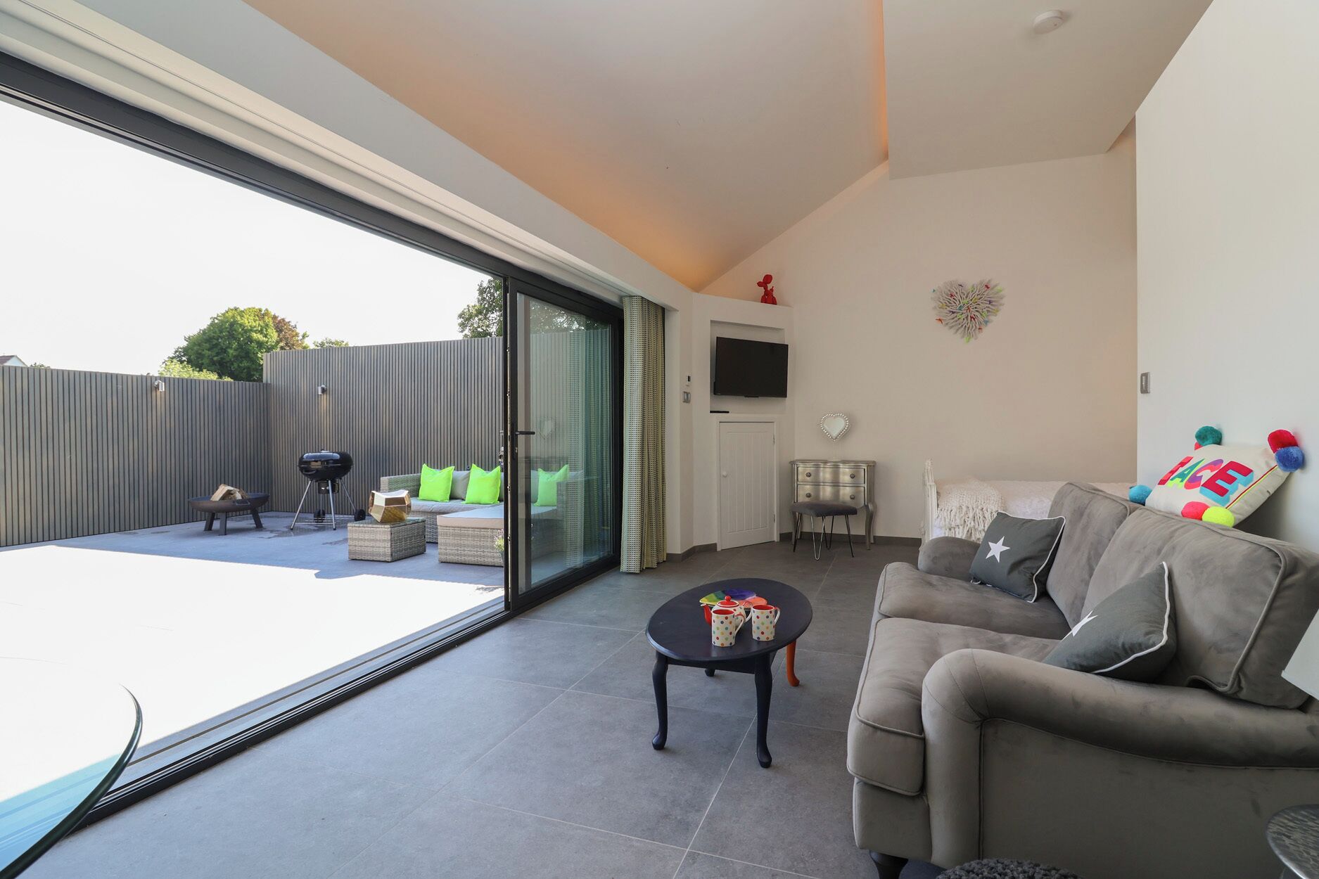 A modern living room with a sofa, a coffee table, and a TV. There is a sliding glass door leading to a patio area with outdoor furniture.