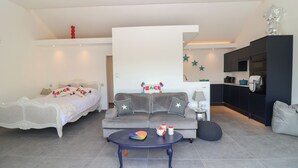 A modern living room with a bed, sofa, and coffee table. There is a kitchen area with cabinets and a refrigerator.