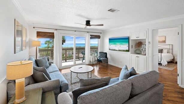 Un vaste espace de vie comprenant un grand canapé, une table basse et un ventilateur de plafond. Un balcon offre une vue sur l’océan.