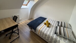 A small, compact room with a bed, a desk, and a chair. There is a skylight above the bed and a window on the left wall. - Apartment in Ecommoy Near Le Mans Circuit (Ecommoy)