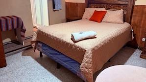 A bedroom with a bed, a wooden headboard, a window with a view, a small table with a striped tablecloth, and a wall-mounted air conditioner.