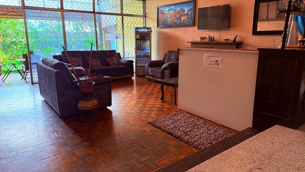 A spacious living area with a sofa, armchairs, a coffee table, and a refrigerator. Large windows provide a view of greenery outside.