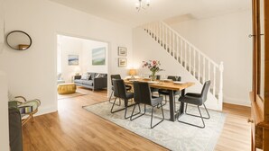 Een eetgedeelte met een houten tafel, zwarte stoelen en een kroonluchter. Een trap met witte leuningen leidt naar een andere kamer.