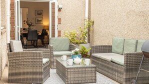Een terras met rieten meubilair, een tafel met kopjes en een theepot, en een deur die naar een binnenruimte leidt.