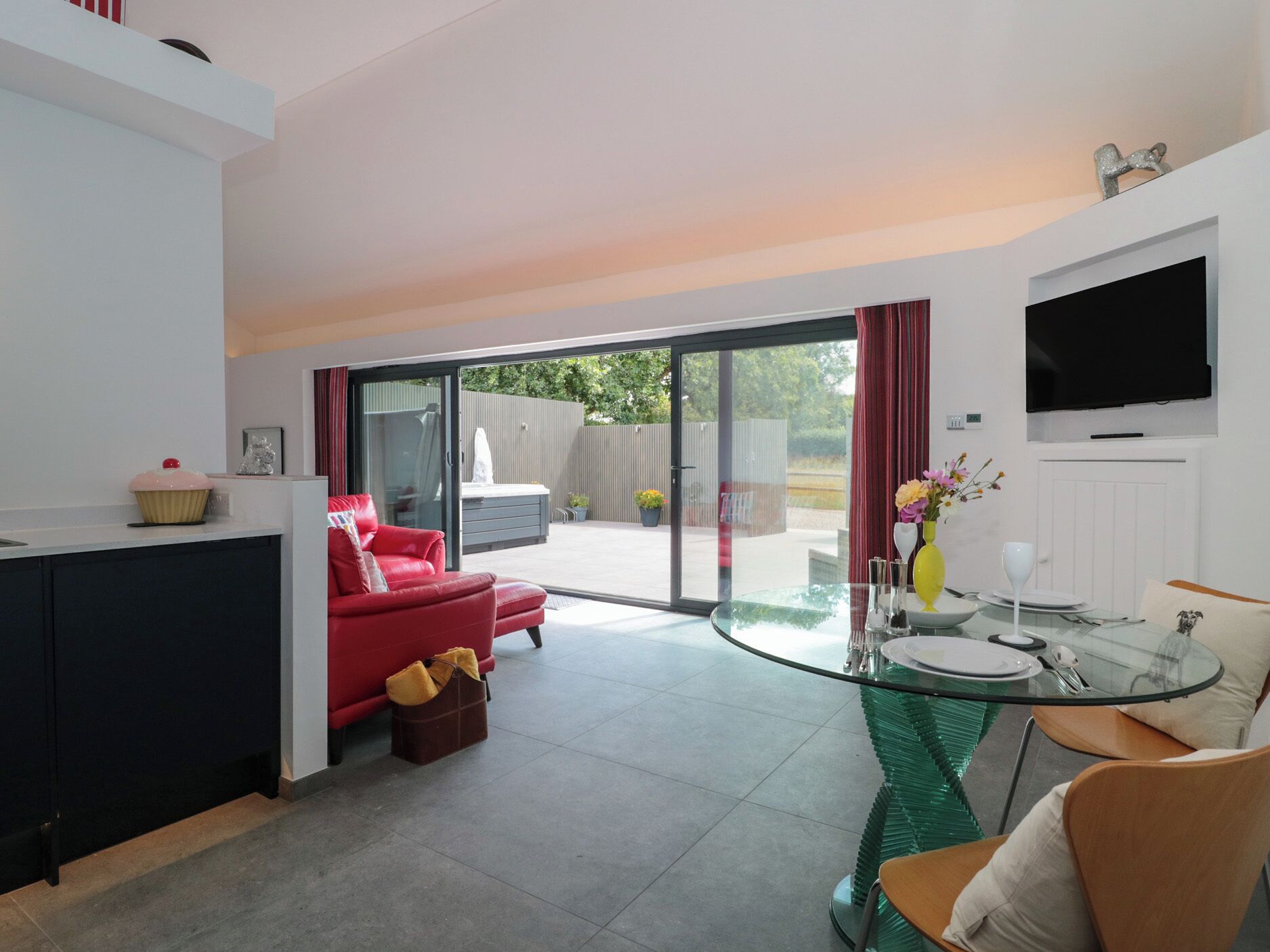 A modern living room with a glass dining table, a red sofa, and a kitchen area with a countertop and a television mounted on the wall.