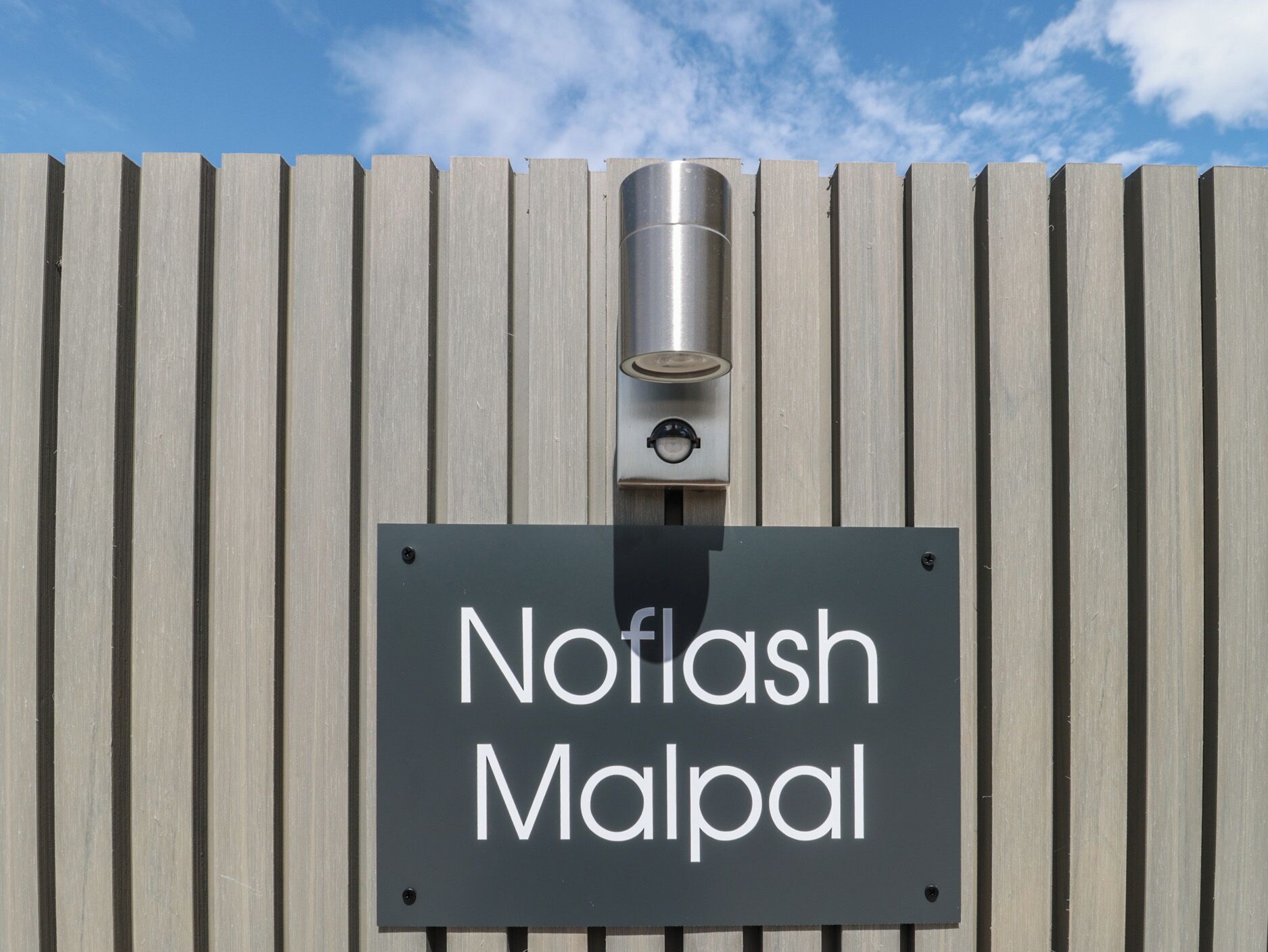 A sign with the name 'Noflash Malpal' mounted on a wall with vertical siding.