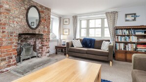 A living room with a brick fireplace, a sofa, a coffee table, a bookshelf, and a window with curtains. - Shelstone (Whitby)