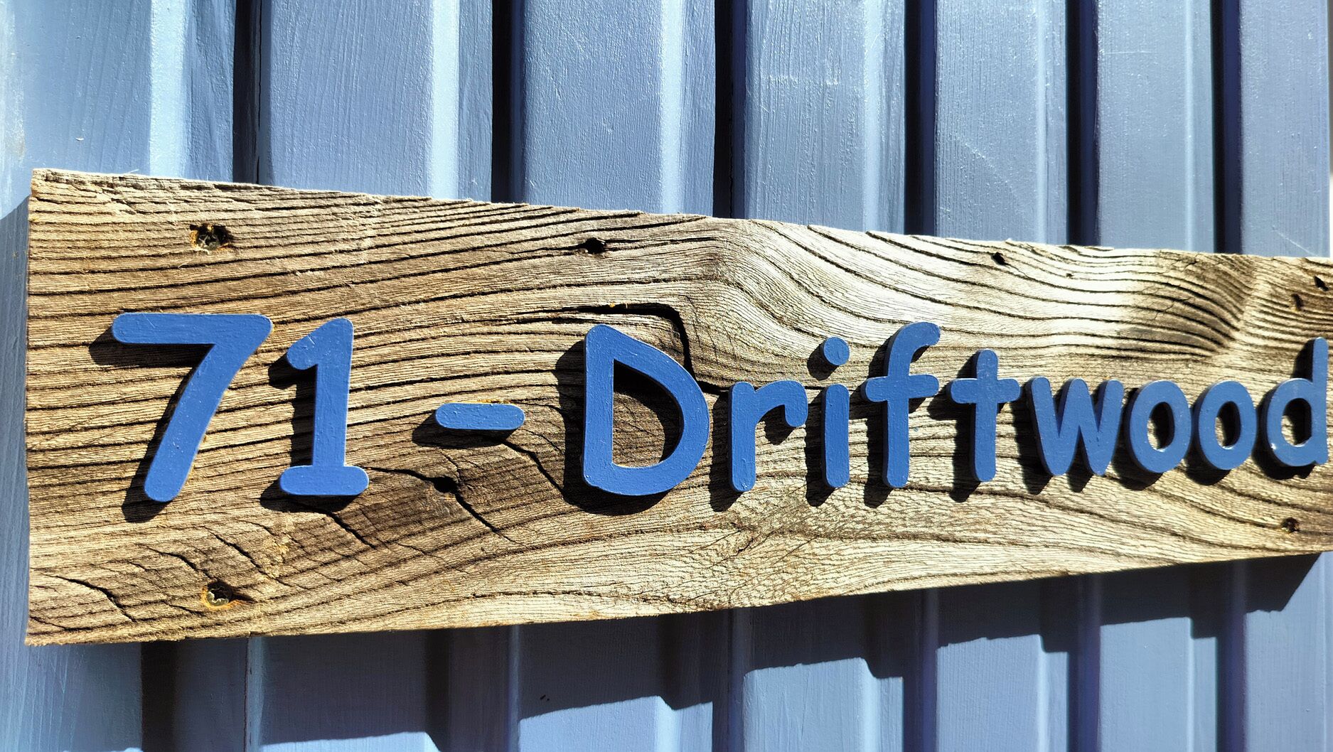 Ein Holzschild mit dem Schriftzug "71-Driftwood" vor einem welligen Metallhintergrund.