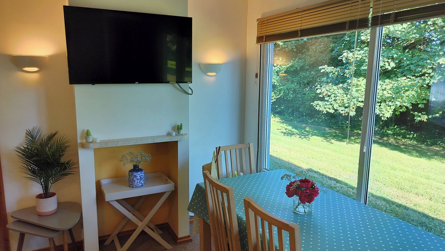 A dining area with a table set for two, a TV mounted on the wall, and a view of a garden through large windows.