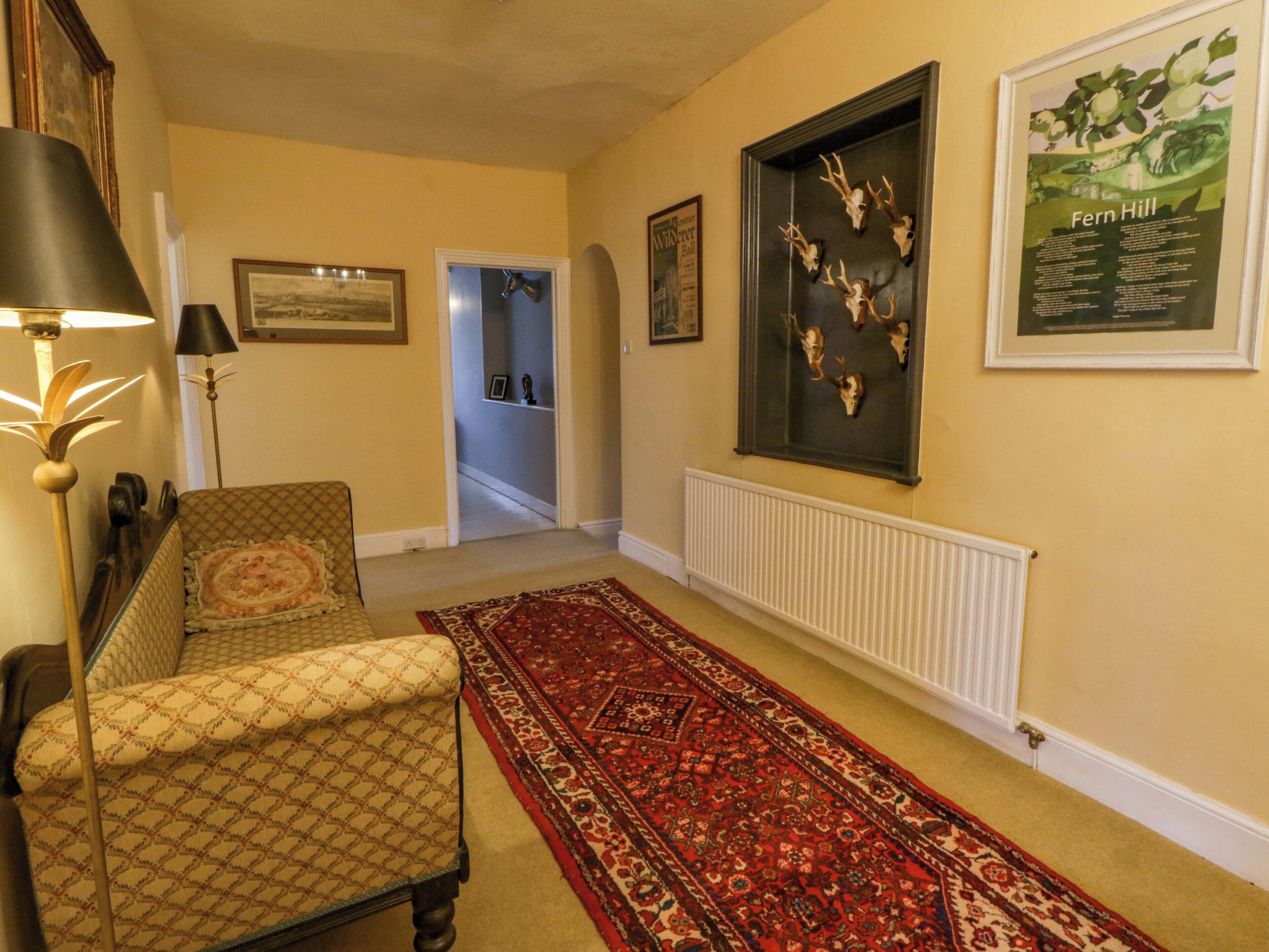 A hallway with a patterned armchair, a red carpet, wall-mounted deer skulls, and a framed poster of Fern Hill.