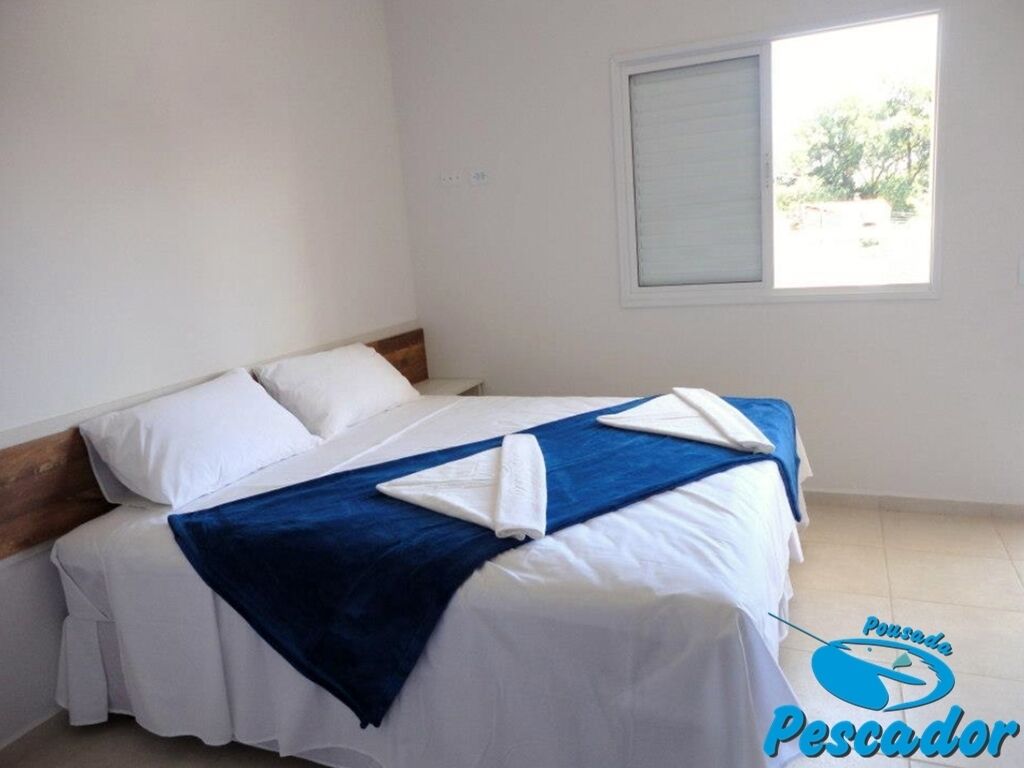 Una cama doble con ropa de cama blanca y azul, una ventana con persianas y un logotipo en la esquina inferior derecha.