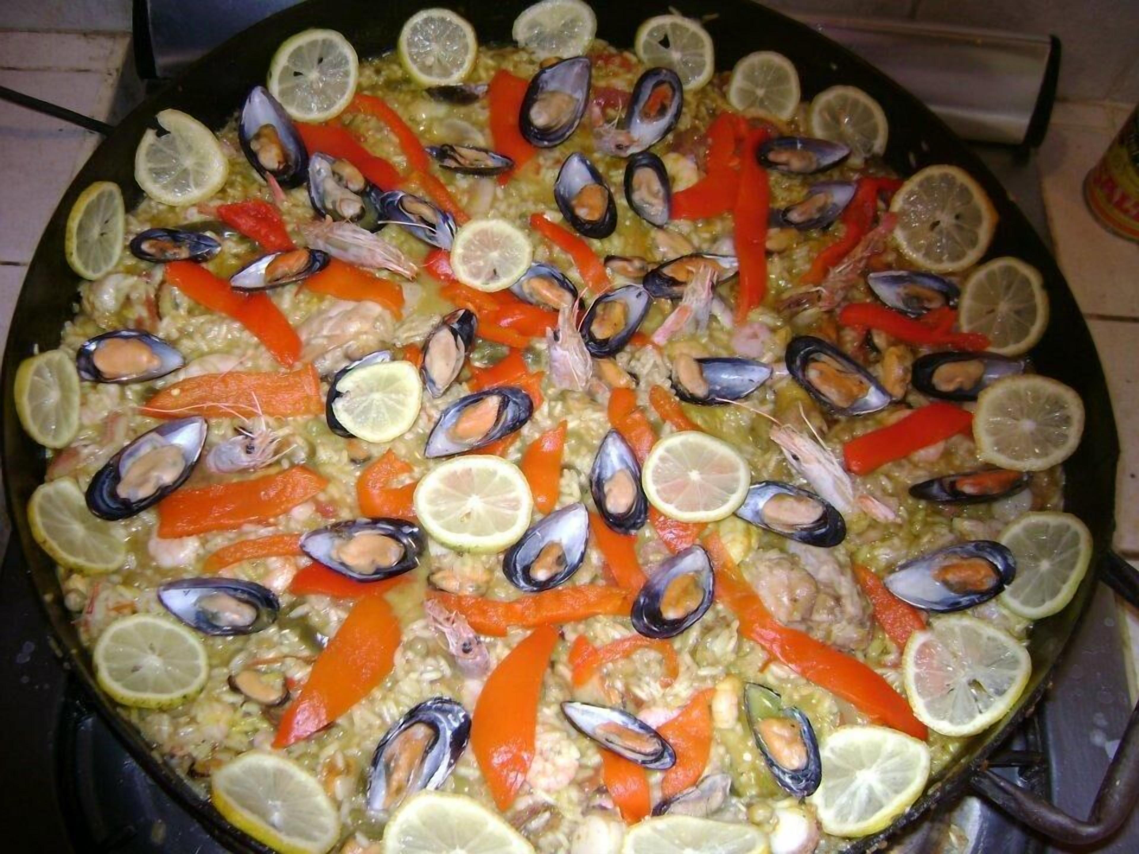 A large, round, shallow cooking pan filled with seafood, including mussels and shrimp, and slices of red bell pepper and lemon.