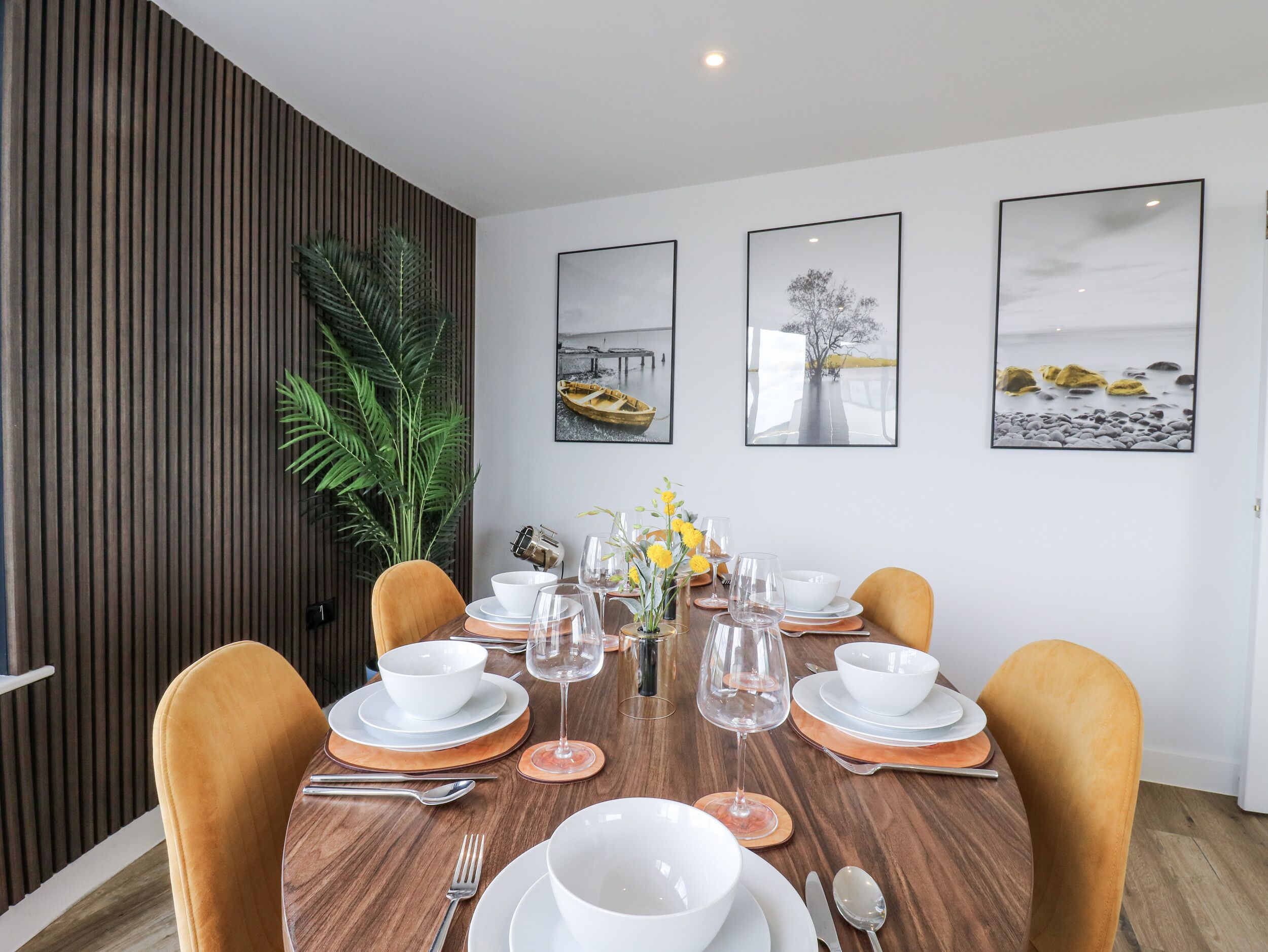 Un comedor con una mesa de madera dispuesta para cuatro, rodeada de sillas. Hay cuadros en la pared, una planta en maceta y una planta verde grande a la izquierda.
