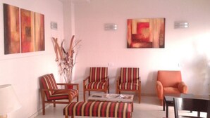 Ein Wohnzimmer mit gestreiften Sesseln, einem Sofa und einem Couchtisch. An den Wänden hängen abstrakte Gemälde, und eine Vase mit Trockenpflanzen steht darauf.