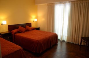 Habitación de hotel con dos camas, cabecero de madera, lámparas de noche y vista al exterior a través de una ventana con cortinas.