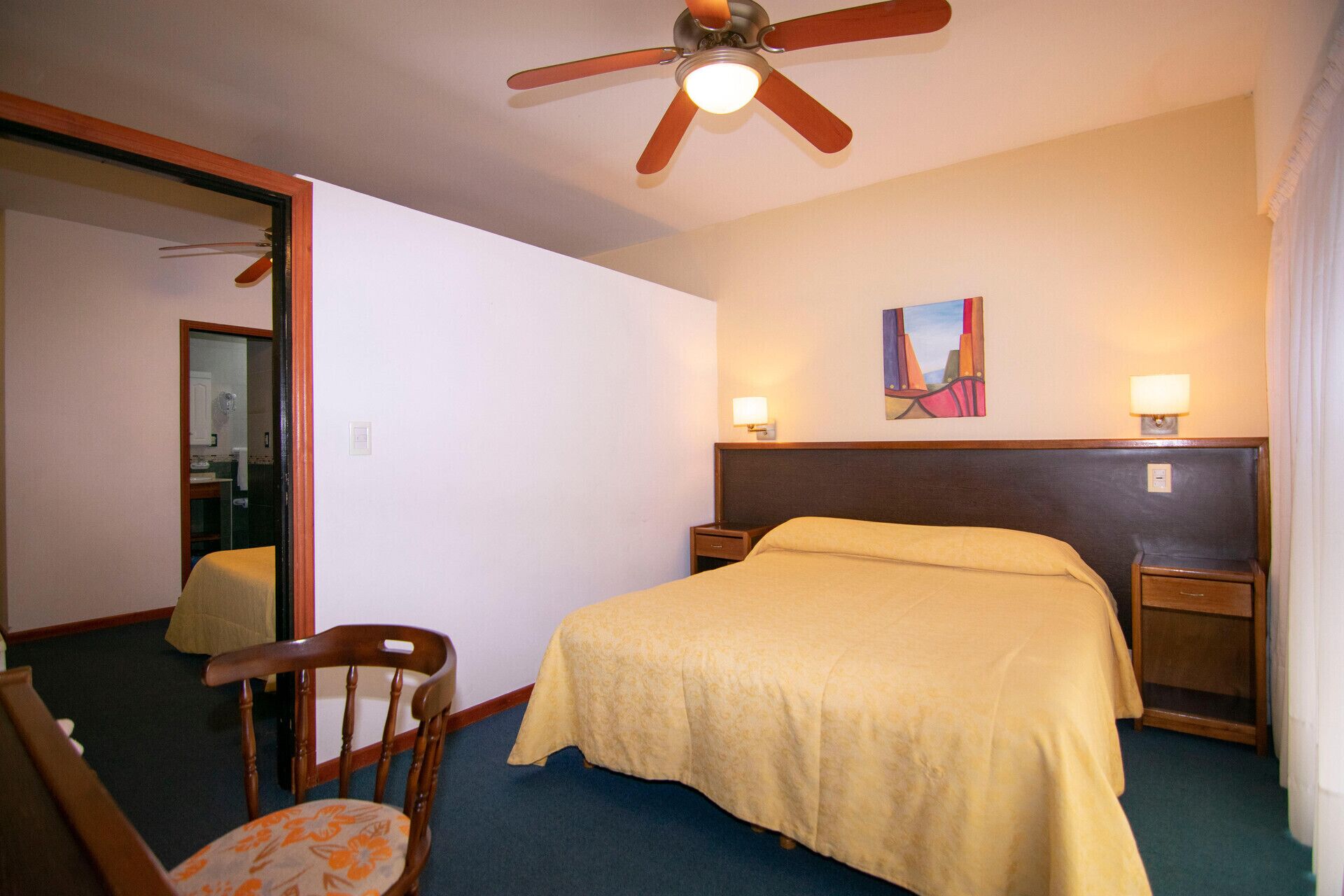 A hotel room with a bed, a chair, a ceiling fan, and a painting on the wall.