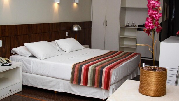Quarto de hotel com cama, luminária de cabeceira, guarda-roupa e uma planta decorativa.