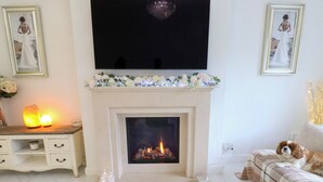 A cozy living room with a fireplace, a sofa, a TV, and decorative elements like framed pictures and candles. - St Mary's House (Oldham)