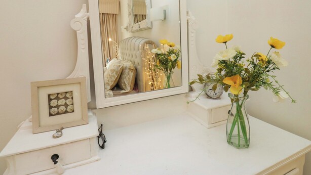 A bathroom vanity with a mirror, a framed picture of roses, a vase with yellow flowers, and a clock. - St Mary's House (Oldham)