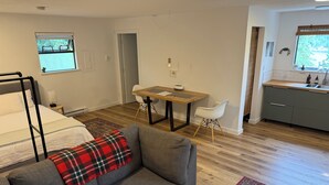 Une pièce moderne et bien éclairée, avec un canapé gris, une table à manger en bois, une petite chaise blanche et un coin cuisine comprenant un évier et des placards.