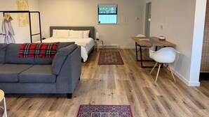 Une pièce moderne et bien éclairée, avec un canapé gris, un lit recouvert de linge de maison blanc, un bureau en bois et une petite table.