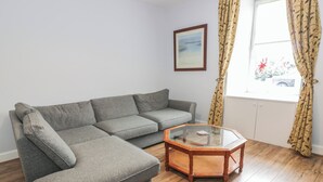 A grey sectional sofa, a glass-top coffee table, wooden flooring, a window with curtains, and a framed picture on the wall. - King's Apartment (Castle Douglas)