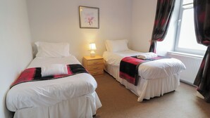 A hotel room with two single beds, a nightstand with a lamp, a framed picture on the wall, and a window with curtains. - King's Apartment (Castle Douglas)